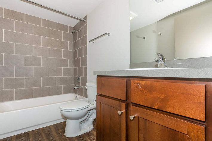 A modern bathroom featuring a gray tiled wall, a white bathtub, a toilet, and a wooden vanity with a gray countertop. Natural light fills the space, creating a bright and clean atmosphere. A large mirror is mounted above the sink, enhancing the sense of openness.