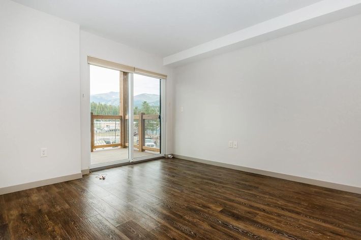 Empty room with a wooden floor, featuring a large sliding glass door that opens to a balcony. Natural light fills the space, highlighting the clean, minimalist design. Mountains are visible in the background through the door. The walls are painted light colors, creating a bright and airy atmosphere.