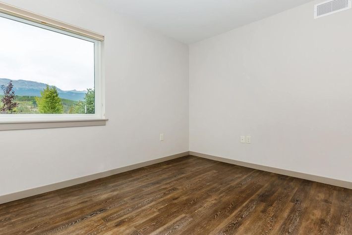 A spacious, empty room with light gray walls and a large window overlooking green trees and distant mountains. The floor is covered in dark wood laminate. The room has no furniture, creating a minimalist and open atmosphere.