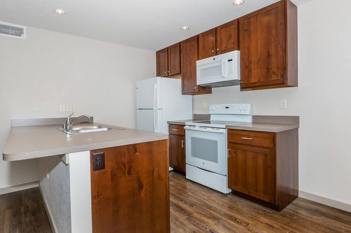 A modern kitchen featuring wooden cabinets, a white stove, microwave, and refrigerator. The countertop is light gray, with a sink on the side. The flooring appears to be wood-like, and the walls are painted a neutral color, creating a bright and inviting space.