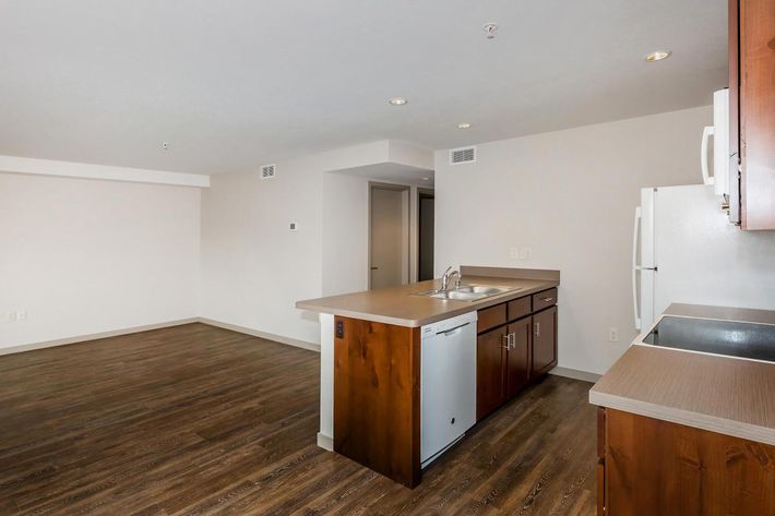 A modern kitchen with a wooden countertop and dark cabinets, featuring a sink and a dishwasher. The open layout includes a spacious living area with light-colored walls and hardwood flooring, enhancing the airy ambiance. A white refrigerator is visible against the wall in the background.