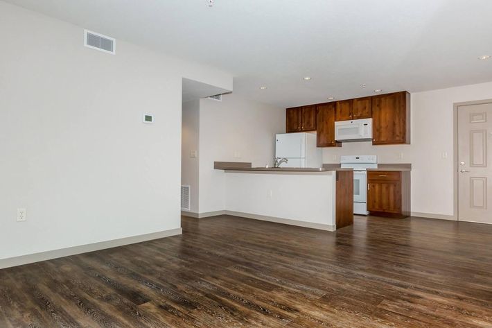 A spacious, modern kitchen and living area featuring wood cabinetry, white appliances, and an open layout. The floor is dark wood, with a light-colored wall. There’s a door leading to another room, and a wall-mounted thermostat is visible. The lighting is bright and inviting.