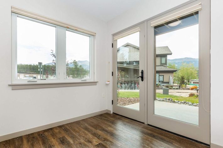 A well-lit room with two large windows and a glass door leading outside. The floor is made of dark wood, and the walls are painted white. Outside, there's a view of a house and mountains in the background, with a landscaped yard visible through the door.