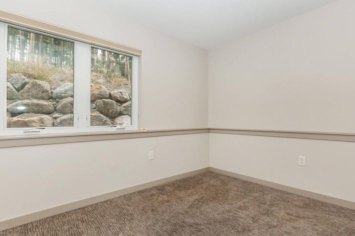 A sparsely furnished room with light beige walls and brown carpeting. There's a window on one wall showing a view of large rocks and greenery outside. The room has a minimalist aesthetic, creating an open and airy feel.
