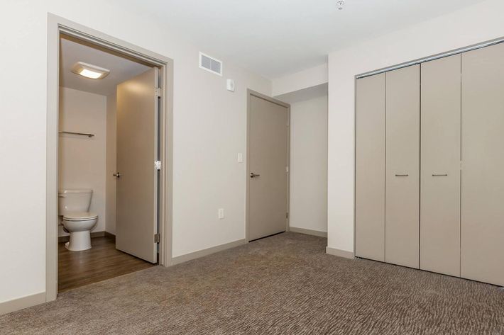 A spacious interior room featuring light-colored walls, carpeted flooring, and two doors leading to different spaces. One door opens to a small bathroom with a toilet, while the other leads to an area with built-in closets. The overall design is modern and neutral.