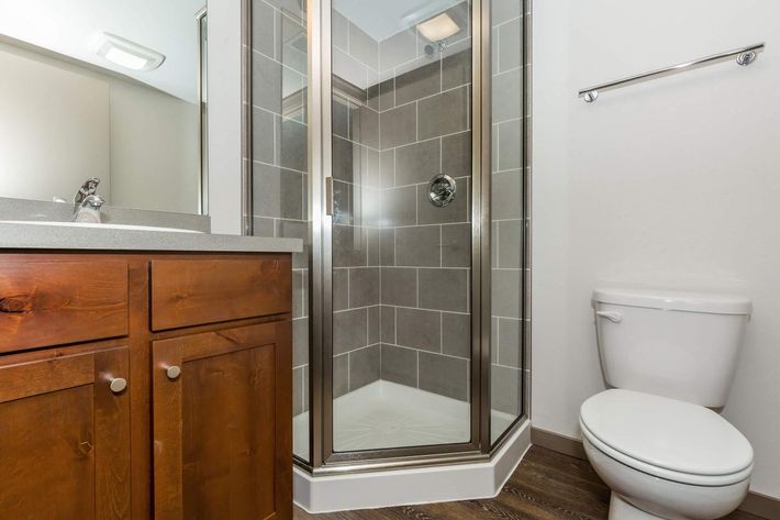 A modern bathroom featuring a glass shower enclosure with tiled walls, a wooden vanity with storage beneath the sink, and a white toilet. The room has neutral-colored walls and a sleek design, creating a clean and functional space.