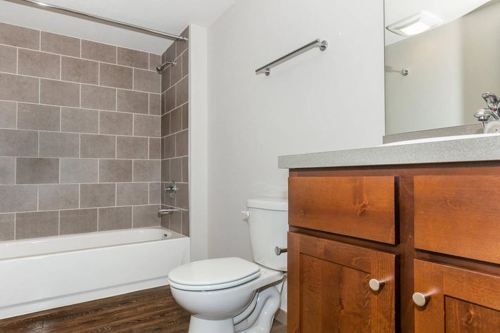 A modern bathroom featuring a bathtub with a showerhead, gray tiled walls, a white toilet, and a wooden vanity with a sink. The floor is styled with dark wood, and there is a wall-mounted mirror and a towel bar. The overall design is clean and contemporary.