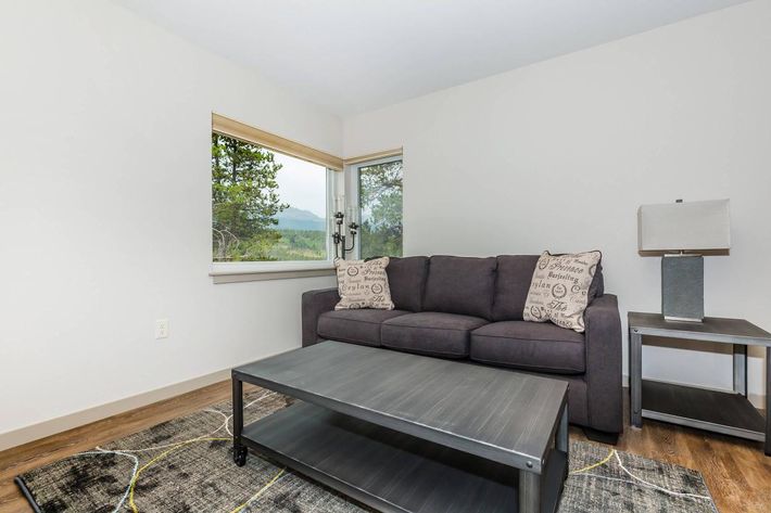 A cozy living room featuring a dark gray sofa with decorative pillows, a sleek coffee table, and a stylish side table with a lamp. A large window offers a view of trees and mountains, allowing natural light to brighten the space. The floor is brown wood, and there is a modern area rug with abstract designs.