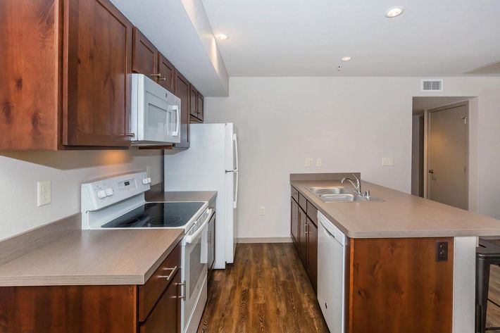 Modern kitchen featuring wooden cabinetry, a white microwave above an electric stove, a refrigerator, and a countertop with a sink. The space has a warm wood floor and neutral wall colors, creating a clean and inviting atmosphere.