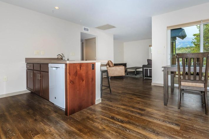 Modern apartment interior featuring an open kitchen with dark wood cabinets, a dishwasher, and a countertop. The living area includes a brown leather chair and a dining table. Large windows provide natural light and a view of outdoor greenery. Hardwood floors complete the contemporary look.