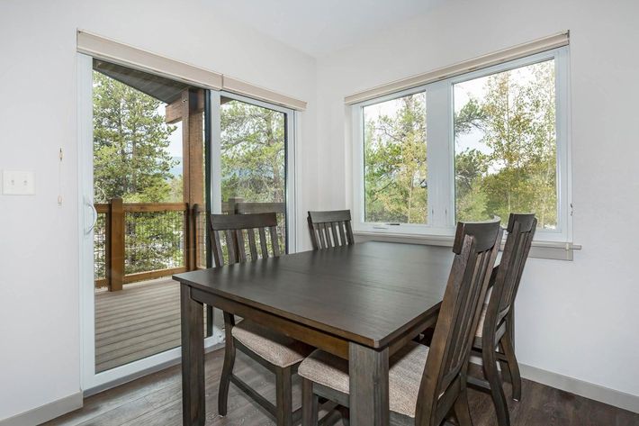 A modern dining area featuring a dark wooden table surrounded by six chairs. Large windows provide a view of trees outside, allowing natural light to fill the space. A sliding door opens to a wooden deck, creating a connection to the outdoors. The walls are painted in a light color, enhancing the bright atmosphere.