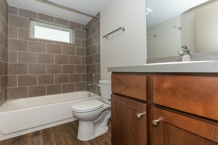 A modern bathroom featuring a tiled wall with brown tones, a white bathtub, a toilet, and a wooden vanity with a sink. The room has a window allowing natural light, and the floor is finished with a wood-like material, creating a clean and contemporary look.