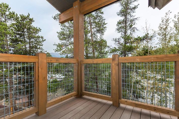 A wooden balcony with a metal mesh railing, surrounded by tall trees. The surface is made of wooden planks, and there are distant views of structures or vehicles partially obscured by foliage. The sky is cloudy, suggesting a serene, outdoor environment.