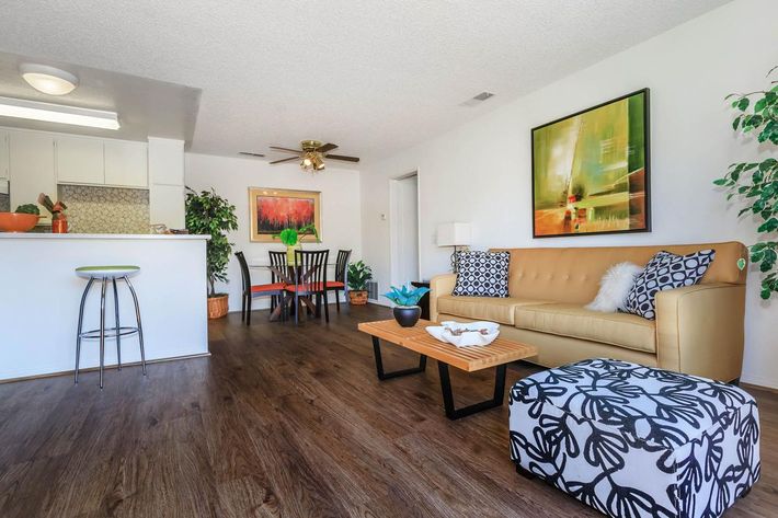 A bright and modern living space featuring a tan couch with pillows, a coffee table, and a patterned pouf. The room includes a dining area with a table and chairs, a well-lit kitchen with bar seating, and decorative plants. Artwork adorns the walls, enhancing the contemporary aesthetic.