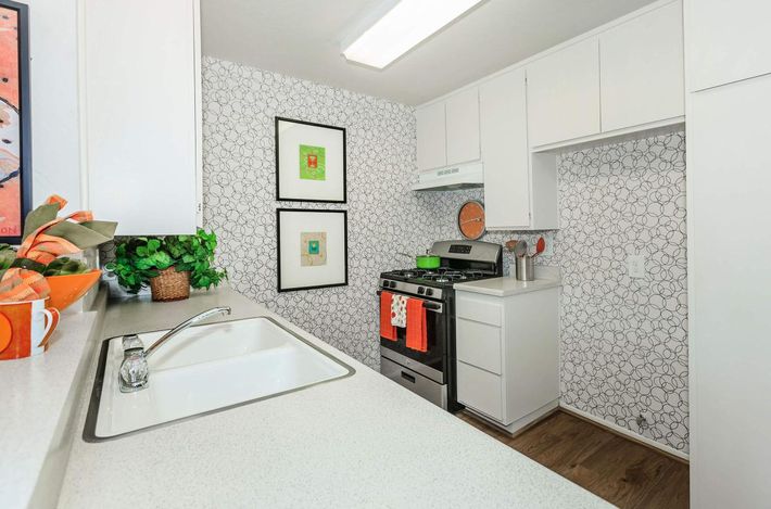 A modern kitchen featuring white cabinetry, a gas stove with an orange clock above, and a sink with a stylish faucet. The walls are decorated with a patterned wallpaper, and there are framed artworks. A potted plant and colorful kitchenware add a vibrant touch to the space.