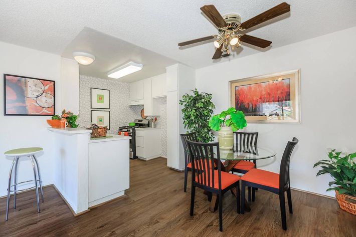 A bright and modern dining area featuring a round glass table with black chairs and orange cushions. The kitchen in the background is partially visible with white cabinetry. Decor includes a potted plant and wall art. A ceiling fan adds a cozy touch to the space.
