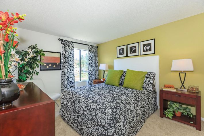 A cozy bedroom featuring a queen-sized bed with patterned bedding and bright green pillows. The walls are painted a warm yellow, and there are decorative curtains. A bedside table with a lamp and plants, along with a dresser holding a vase of flowers, complete the inviting ambiance. Large windows allow natural light to enter.