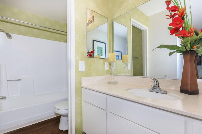 A modern bathroom featuring a white bathtub and shower combination, a white vanity with a sink, and a green accent wall. The space is decorated with a floral arrangement in a tall vase, candles, and framed artwork on the walls, creating a bright and inviting atmosphere.