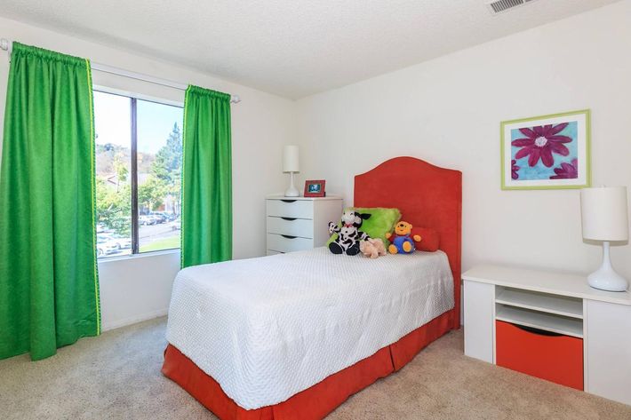 A bright and cheerful children's bedroom featuring a twin bed covered with a white quilt and adorned with colorful stuffed animals. The walls are painted in neutral tones, complemented by vibrant green curtains and an orange headboard. A small dresser and a shelf with decorative items complete the cozy space, with a view outside the window.