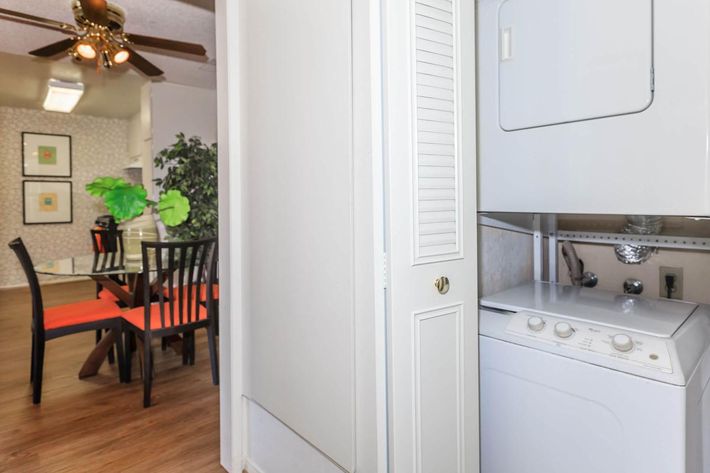 A laundry area featuring a stacked washer and dryer, with a closet door beside it. In the background, there's a dining area with a wooden table and black chairs, accented by red cushions, and a ceiling fan overhead. The walls are decorated with framed artwork and a potted plant adds a touch of greenery.