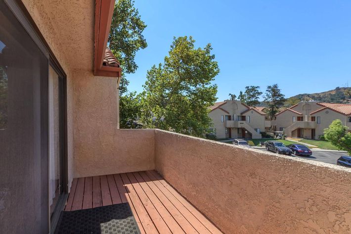 A spacious balcony with wooden flooring and a textured mat, offering a view of neighboring houses and trees under a clear blue sky. The scene captures the peaceful exterior of a residential area, with parked cars visible in the distance.