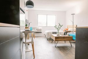 A bright, modern studio apartment featuring a small dining table with a white chair, a cozy bed with a wooden headboard, a blue couch, and a wooden coffee table. The space is well-lit with a large window and has minimalistic decor, including a floor lamp and potted plants. Air conditioning unit visible on the wall.