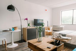 A modern, minimalist studio apartment featuring a bed, a flat-screen TV on a wooden cabinet, a round dining table with two chairs, a tall floor lamp, and decorative plants. A large window lets in natural light, and a small air conditioning unit is visible. The decor is simple and inviting.
