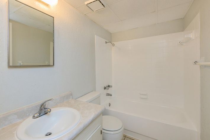A clean and simple bathroom featuring a white tub and shower combination, a sink with a countertop, and a mirror. The walls are painted light grey, and the floor is tiled. The bathroom has a bright ceiling light and basic fixtures, creating an inviting and functional space.