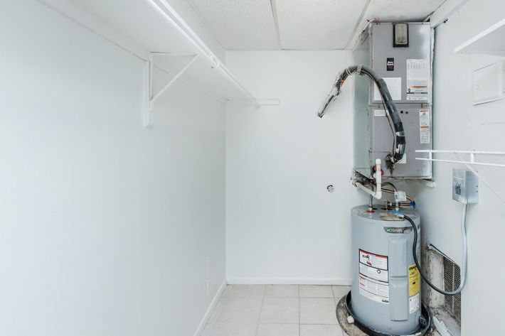 A clean, empty laundry room featuring white walls and a simple tile floor. On the right, there is a water heater connected to pipes. There are also shelves mounted on the wall for storage. The space is bright and well-lit, creating an airy atmosphere.