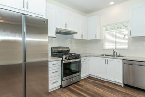 Modern kitchen featuring stainless steel appliances, including a large refrigerator and a gas stove with an oven. The cabinetry is white with silver handles, and there is a sink situated beneath a window that lets in natural light. The floor has a warm wood finish, creating a welcoming atmosphere.