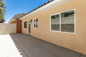 Side view of a single-story residential house featuring a beige stucco exterior, multiple windows with white frames, and a concrete patio. A white fence provides privacy, and clear blue skies are visible above, creating a bright and open atmosphere.