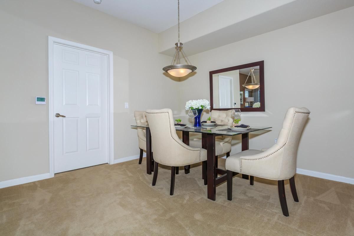 A well-lit dining area featuring a glass table surrounded by four beige upholstered chairs. A vase of flowers sits in the center, and a decorative mirror is mounted on the wall. The walls are painted a neutral color, and there is a door visible in the background, adding to the cozy ambiance.