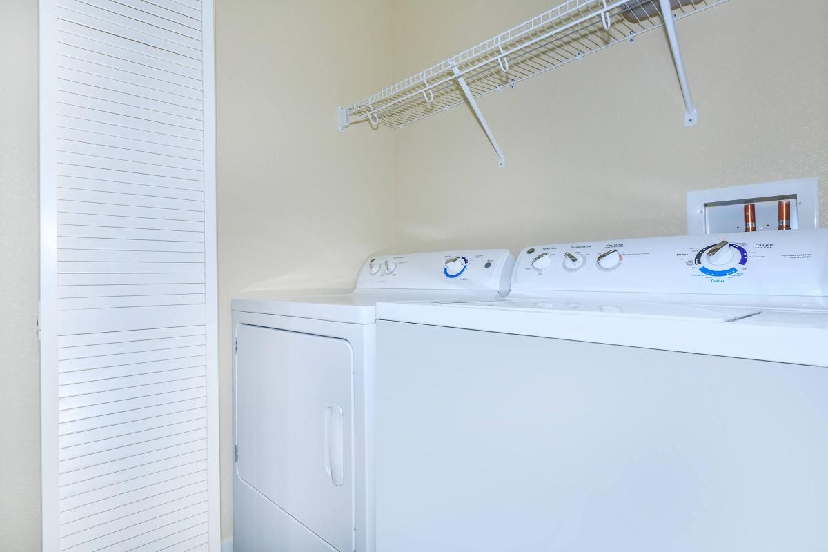 A laundry room featuring a stacked washer and dryer unit against a light-colored wall. Above the appliances, there is a wire shelving unit for storage. A closed white bi-fold door is visible on the left side of the image, providing access to additional storage space.