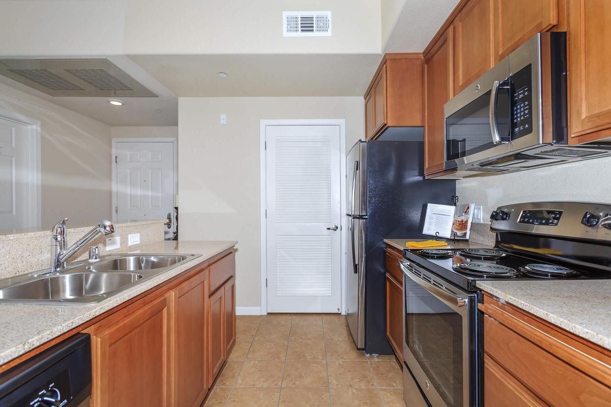 Modern kitchen featuring wooden cabinets, stainless steel appliances including an oven, microwave, and refrigerator. A double sink is positioned near the window, with tiled flooring and light-colored walls, creating a bright and inviting atmosphere. A door leads to another room or area.