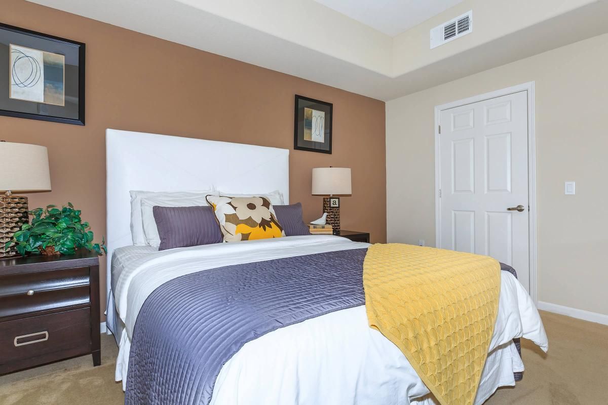 A cozy bedroom featuring a large white bed with a lavender and yellow throw pillow, a grey quilt, and a yellow blanket. Two bedside tables with lamps are placed on either side of the bed. The walls are painted a warm brown, and framed art pieces are hung above the bed and on the wall. A closed door is visible in the background.