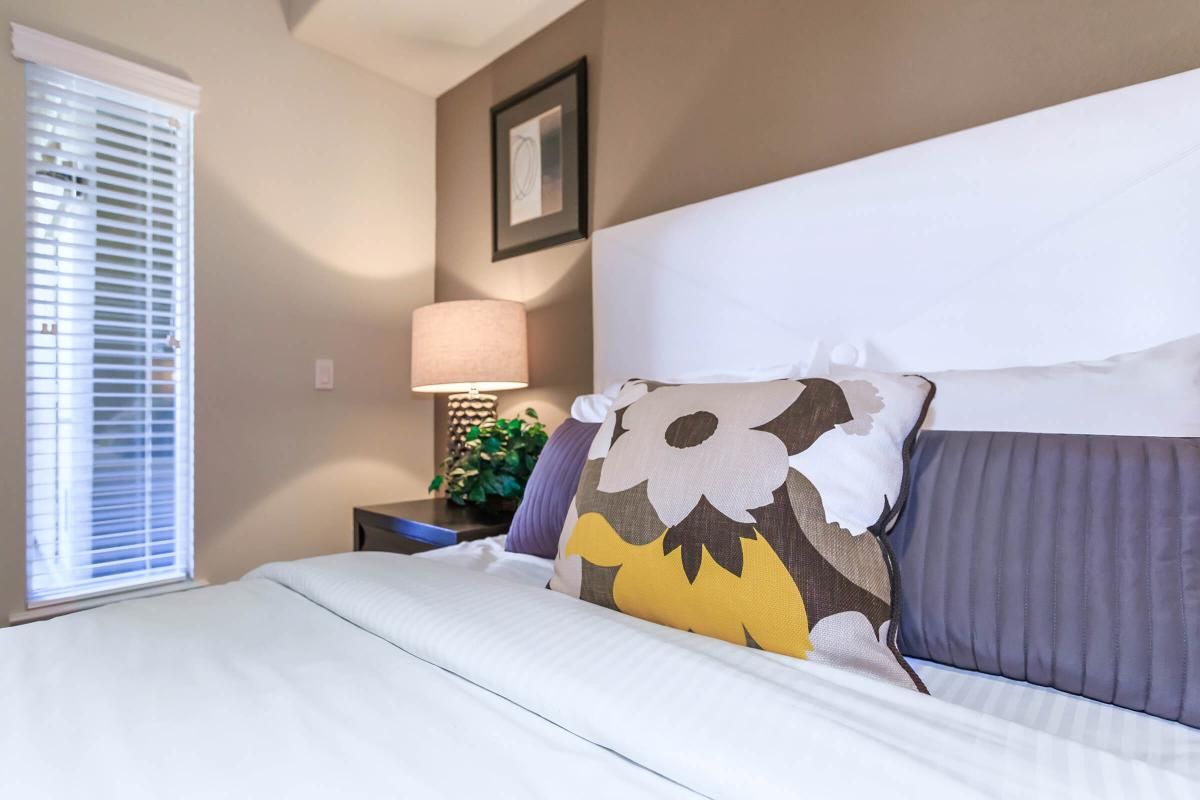 A stylish bedroom featuring a neatly made bed with a decorative floral pillow. There’s a bedside table with a lamp, and a window covered with white blinds, allowing natural light to softly illuminate the room. A small plant adds a touch of greenery to the cozy space.