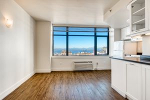 Bright, spacious apartment interior featuring large windows with a panoramic view of mountains and cityscape. The room has light-colored walls and modern wood flooring. A minimalist kitchen area with white cabinets is visible on the right, enhancing the open and airy feel of the space.