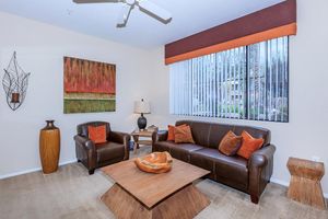 A cozy living room featuring a brown leather sofa with orange accent pillows, a matching armchair, and a wooden coffee table. A decorative vase and lamp are present, along with a large painting on the wall and a window with vertical blinds allowing natural light to filter in.