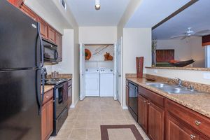 A modern kitchen featuring dark wood cabinetry, stainless steel appliances, and granite countertops. There is a double sink, a stove, and a black refrigerator. In the background, a laundry area with a washer and dryer is visible, along with decorative wall art and plants. The flooring is tiled.