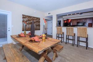 A modern dining area featuring a rustic wooden table set with decorative plates and cloth napkins. In the background, there is a fully equipped kitchen with dark wood cabinetry and bar stools. The walls are adorned with artwork, and the room is well-lit with natural light.