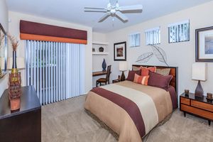 A cozy bedroom featuring a large bed with a patterned beige and brown bedspread. There are decorative pillows in various shades. A ceiling fan hangs above, and the room has a window with vertical blinds. Light beige walls are accented with framed artwork and a small desk with a chair, alongside a nightstand with table lamps.