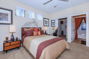 A well-decorated bedroom featuring a queen-sized bed with brown and beige bedding, two bedside lamps on wooden nightstands, and framed artwork on the walls. There are light-colored walls and partially open window blinds, contributing to a bright atmosphere. A walk-in closet is visible on the left, and a bathroom door is slightly ajar on the right, showcasing a neutral tile flooring.