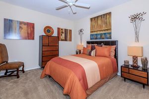 A cozy bedroom with a large bed featuring orange and beige bedding. The room is decorated with abstract wall art and has a wooden dresser and nightstands with lamps. A comfortable chair is positioned in the corner, and the carpet is a light beige color.