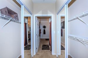A well-lit hallway with two open doorways leading to bathrooms. Each bathroom has a towel rack and a large mirror. There are shelves on the walls and dark rugs on the tiled floor.