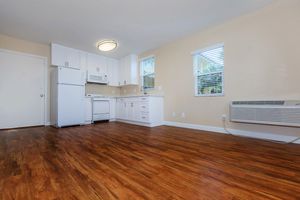 A bright kitchen with light-colored walls, wooden laminate flooring, white cabinets, and modern appliances. It features a refrigerator, oven, and dishwasher, along with a window allowing natural light. An air conditioning unit is visible on the wall. The space appears clean and well-maintained.