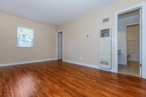 Empty room featuring hardwood flooring, light beige walls, and a small window with blinds. A doorway leads to another room, and there is a bathroom entrance visible on the right. An air conditioning unit is mounted on the wall near the entrance. Natural light brightens the space.