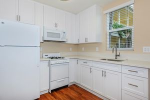 Modern kitchen featuring white cabinets, a white refrigerator, and a microwave above a gas stove. Includes a stainless steel sink under a window, with light-colored countertops and warm wood flooring. Natural light floods the space, creating a bright and inviting atmosphere.