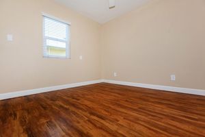 A simple, empty room with light brown walls and a wooden floor. A small window with blinds allows natural light to enter, and there are two wall switches visible. The space is unfurnished, creating a clean and open feel.