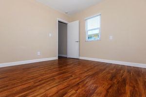 A spacious, empty room with light beige walls and a polished wooden floor. A partially open white door on the left leads to another area, and a window with horizontal blinds on the right allows natural light to illuminate the space. The room appears clean and ready for furnishings.