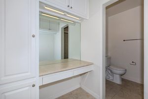 A bright, clean bathroom with a mirrored vanity and lighted cabinetry. The space features a beige countertop and light-colored walls. There is a toilet in a separate nook visible through an open door, enhancing the room's functionality.